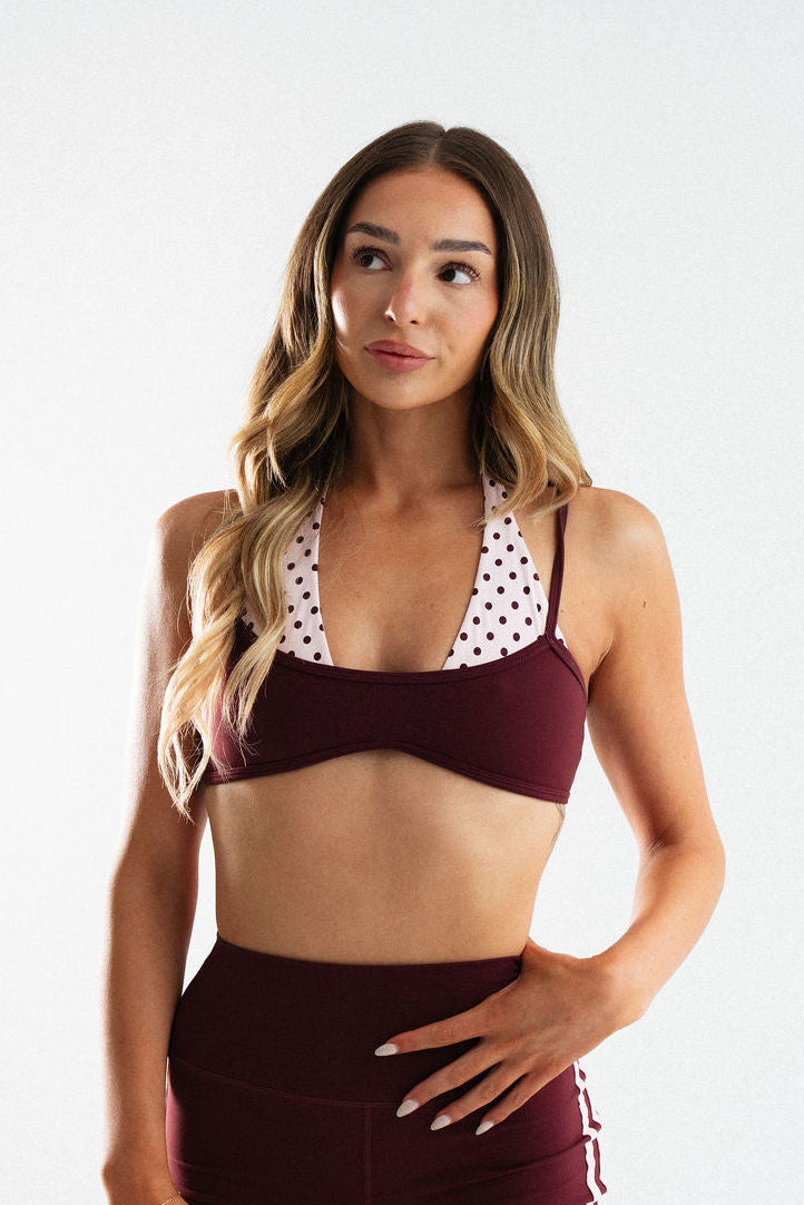 A woman modeling a matching burgundy activewear set consisting of a sports bra with white polka-dot trim and high-waisted shorts with white side stripes, against a light gray studio background.