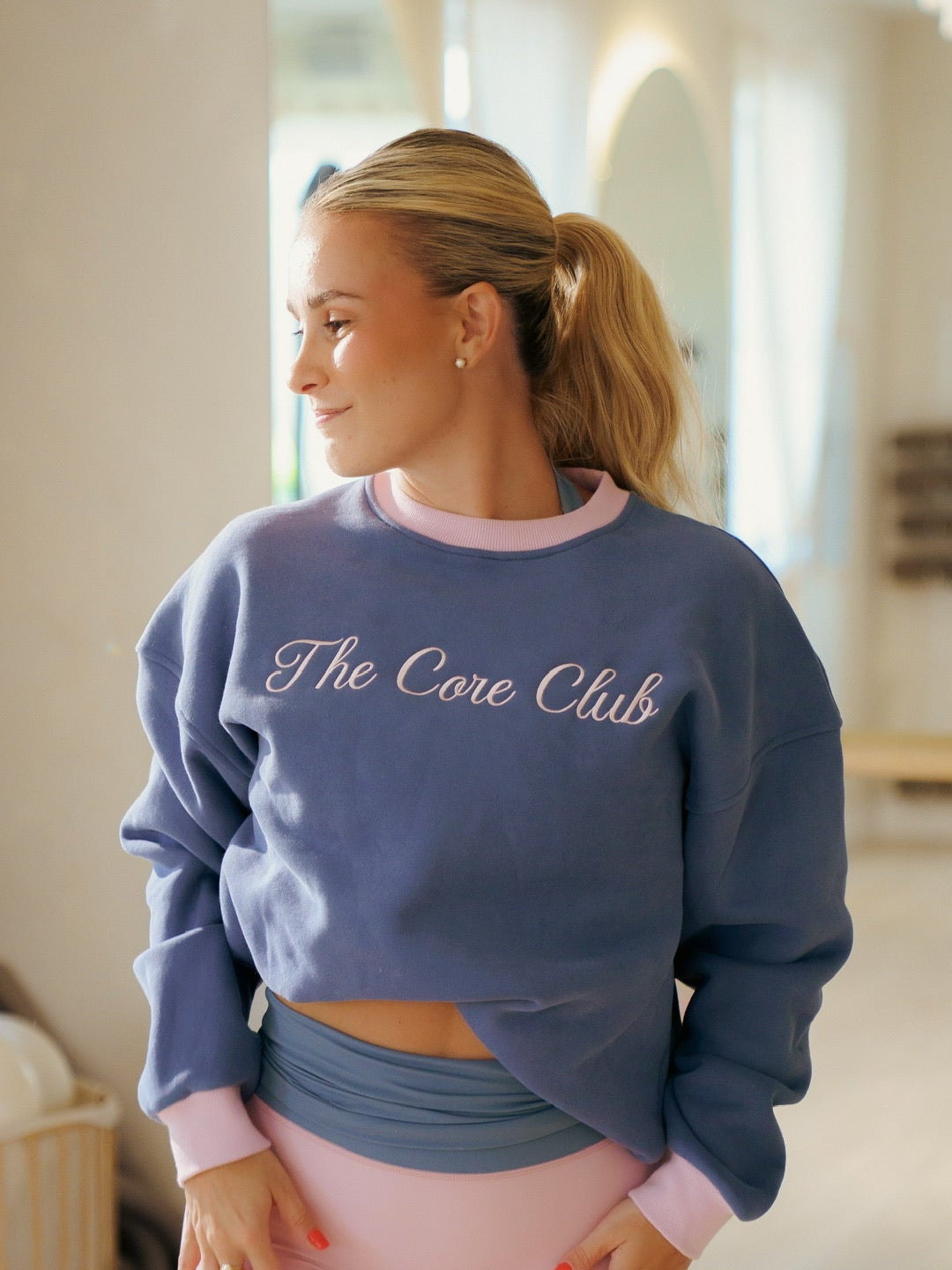 A woman in a gym setting wearing a dusty blue cropped sweatshirt with 'The Core Club' written in white script across the chest, paired with light pink athletic shorts.